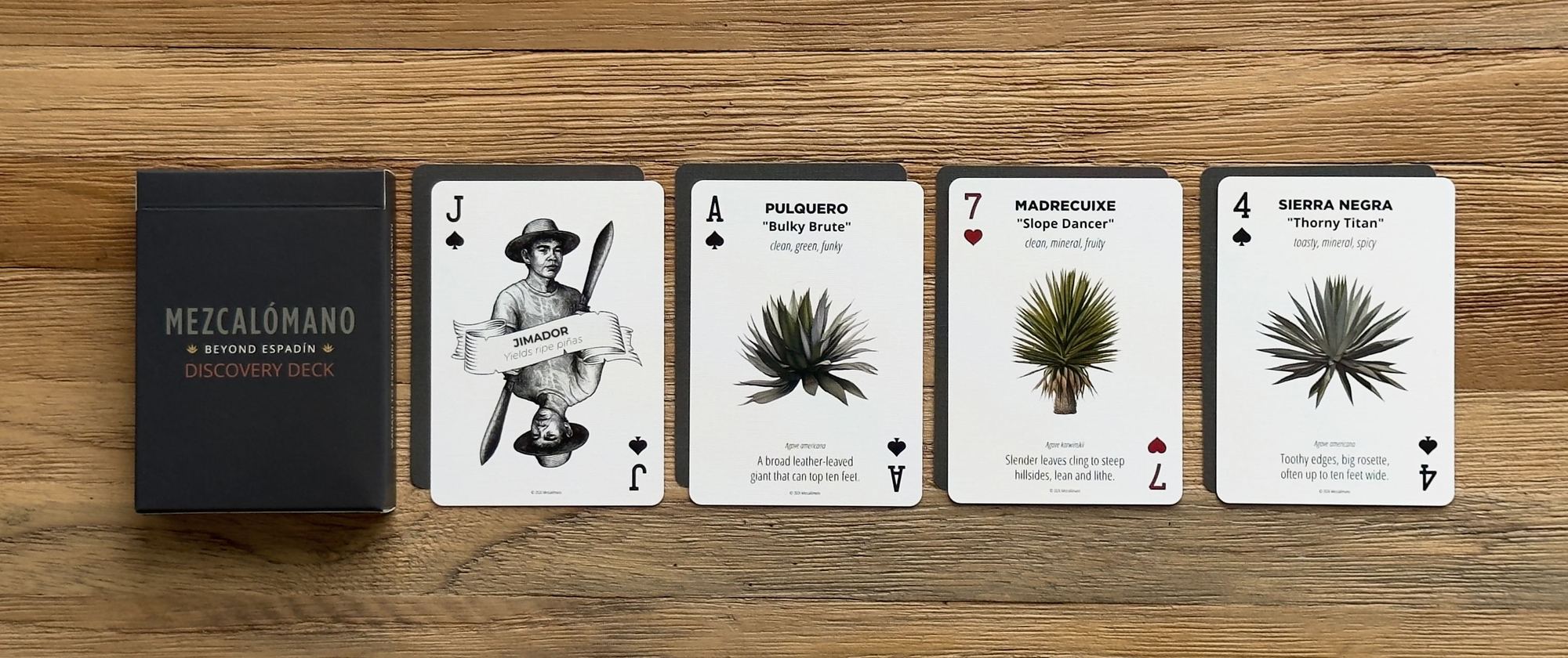The Mezcalómano Discovery Deck box beside four agave cards laid out in a row on a wood table.