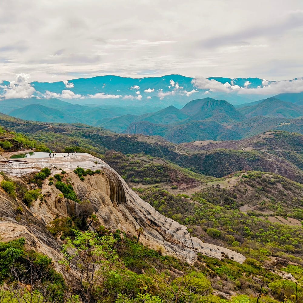The view from Hierve el Agua in Oaxaca, with green valleys and layered mountains under a cloudy sky.